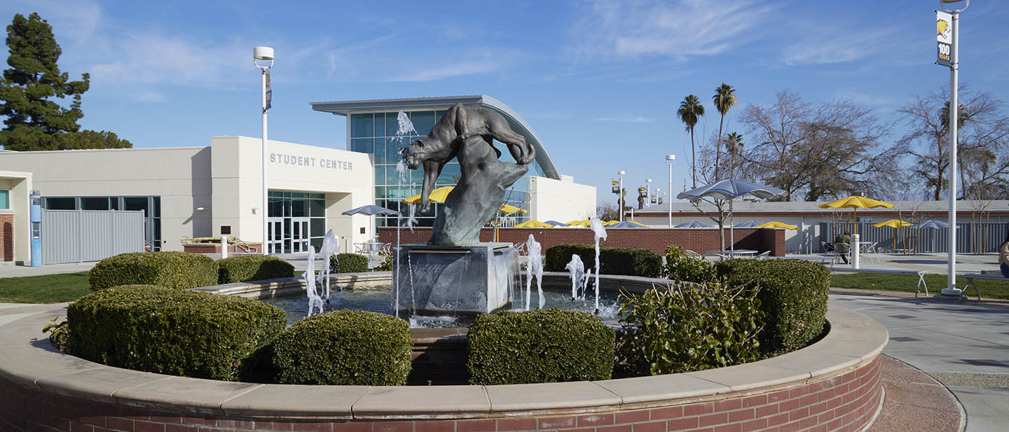 circular fountain with bronze cougar sculpture in the center and water spouts, surrounded by circular low brick wall and shrubs; background is the Student Center building with a tall glazed wall.