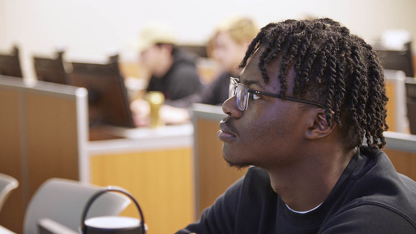 Male student seated at desk in classroom looks toward front of room.