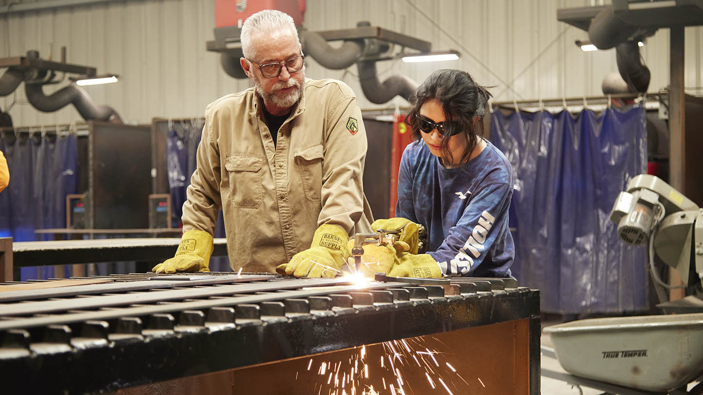 Female high school-aged student in protective face helmet points flame of hand-held welding torch at table and sparks fly out below.