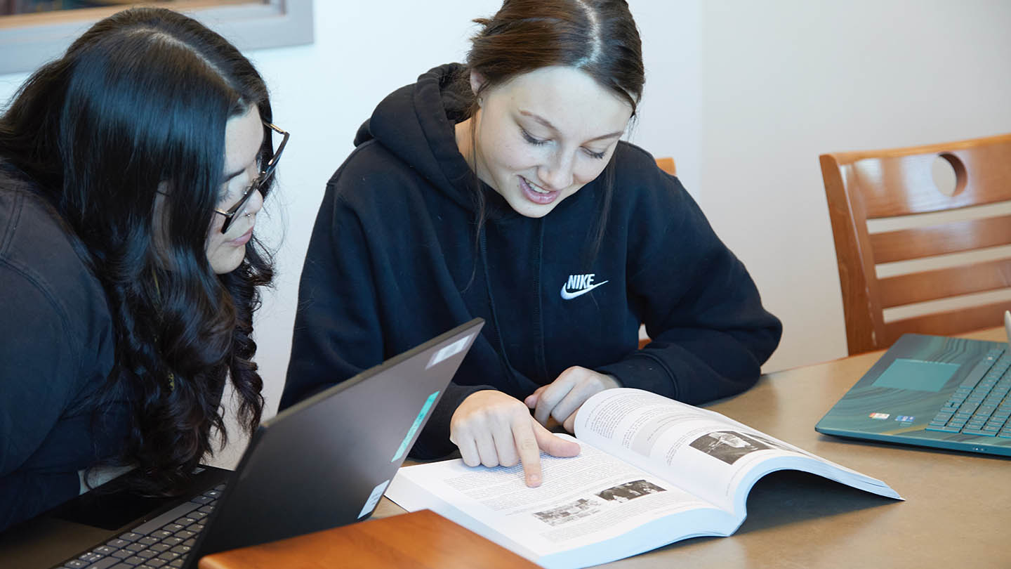 One seated female student points to her textbook as another at her laptop looks over at the book.