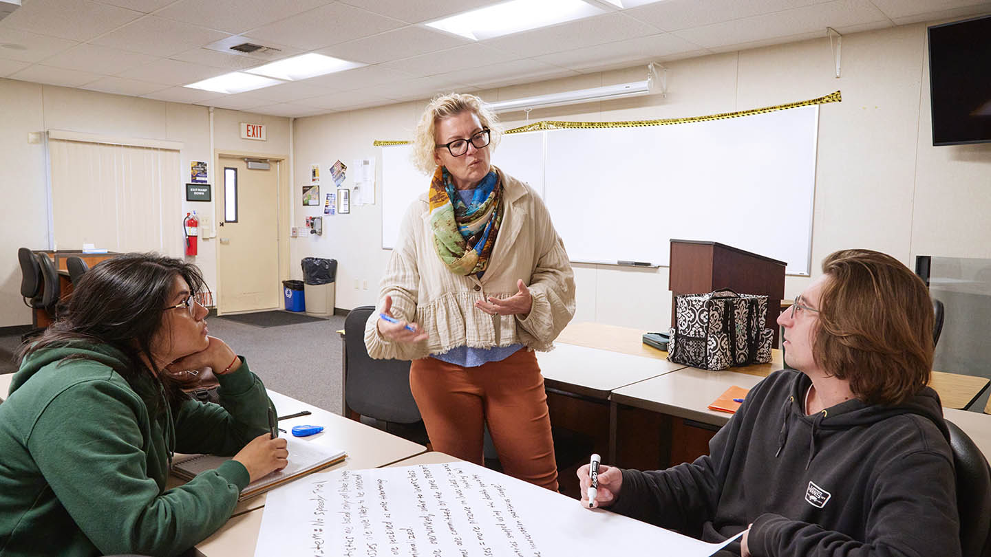 Female professor talks to two students who are writing notes on a poster-sized paper with a thick marker pen.