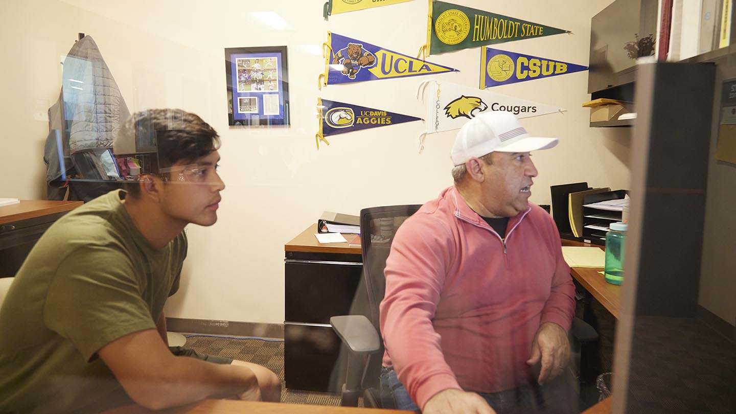 Mature male seated at desk turns to his computer screen on wall and moves the mouse controller as male student concentrates on screen images.