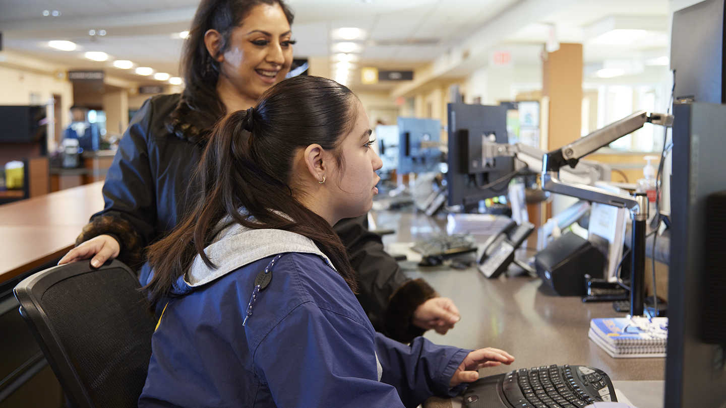 Female coordinator stands next to seated female student looking at a computer screen.