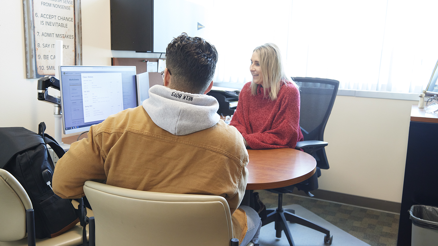 A female counselor sits at a round table with a male, seen from the back, and they look at information on a bright computer screen.