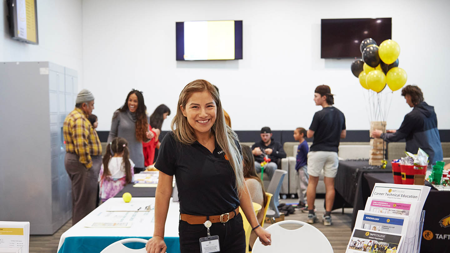 Smiling female at Counseling Table in a room with other tables with exhibits and milling students.